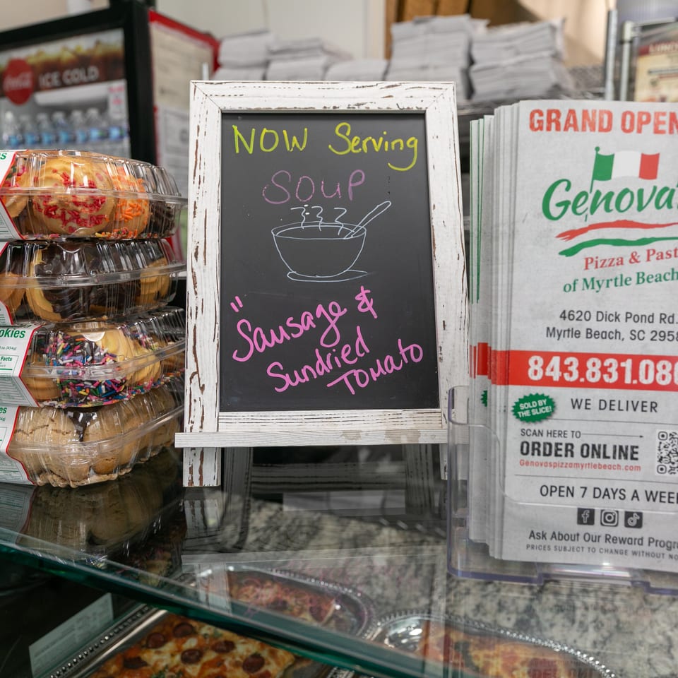 A chalkboard sign advertises "NOW Serving Soup: Sausage & Sundried Tomato" next to Genova's Pizza & Pasta flyers.