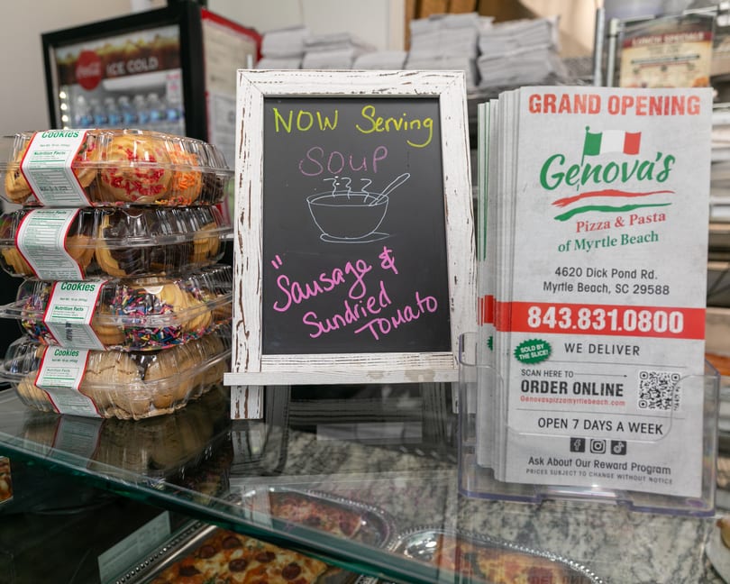 A chalkboard sign advertises "NOW Serving Soup: Sausage & Sundried Tomato" next to Genova's Pizza & Pasta flyers.