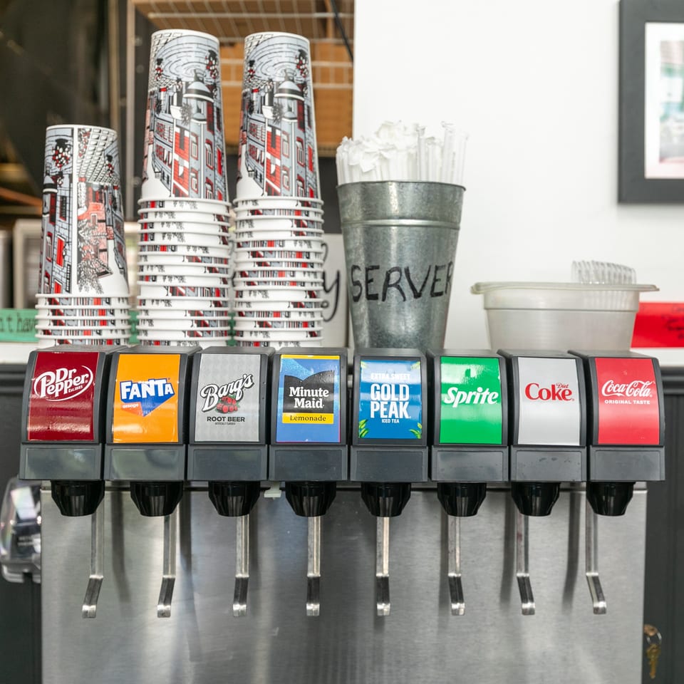 A soda fountain with various drink options, stacked cups, and a "SERVER" bucket for straws.