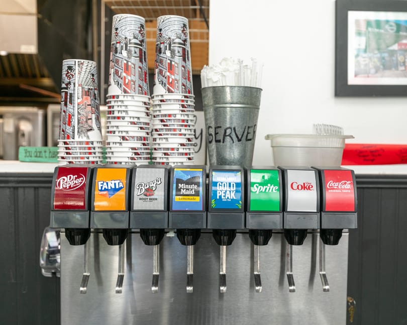 A soda fountain with various drink options, stacked cups, and a "SERVER" bucket for straws.