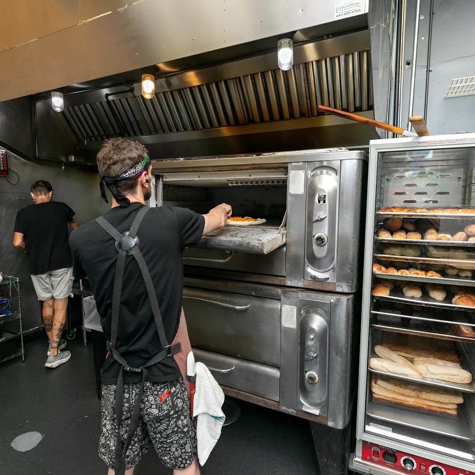 A chef places a pizza into a large oven, with another chef and bread racks nearby.