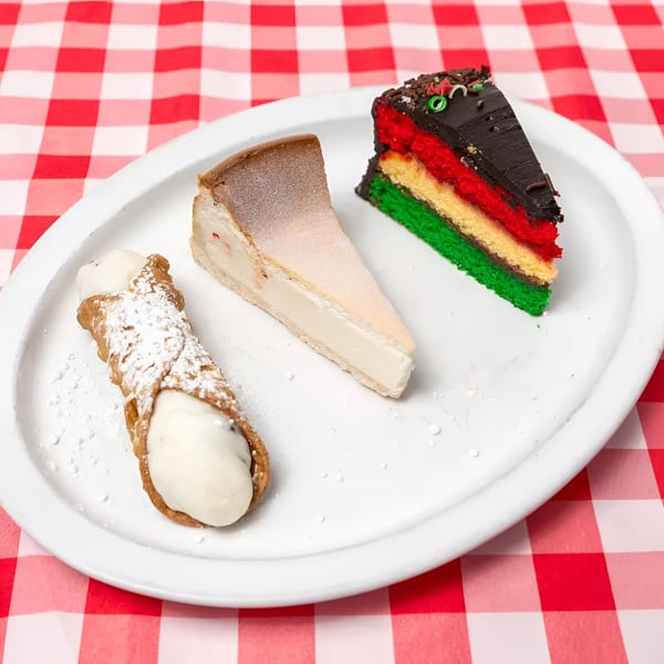 A cannoli, cheesecake slice, and rainbow cake slice on a white plate over a red and white checkered tablecloth.