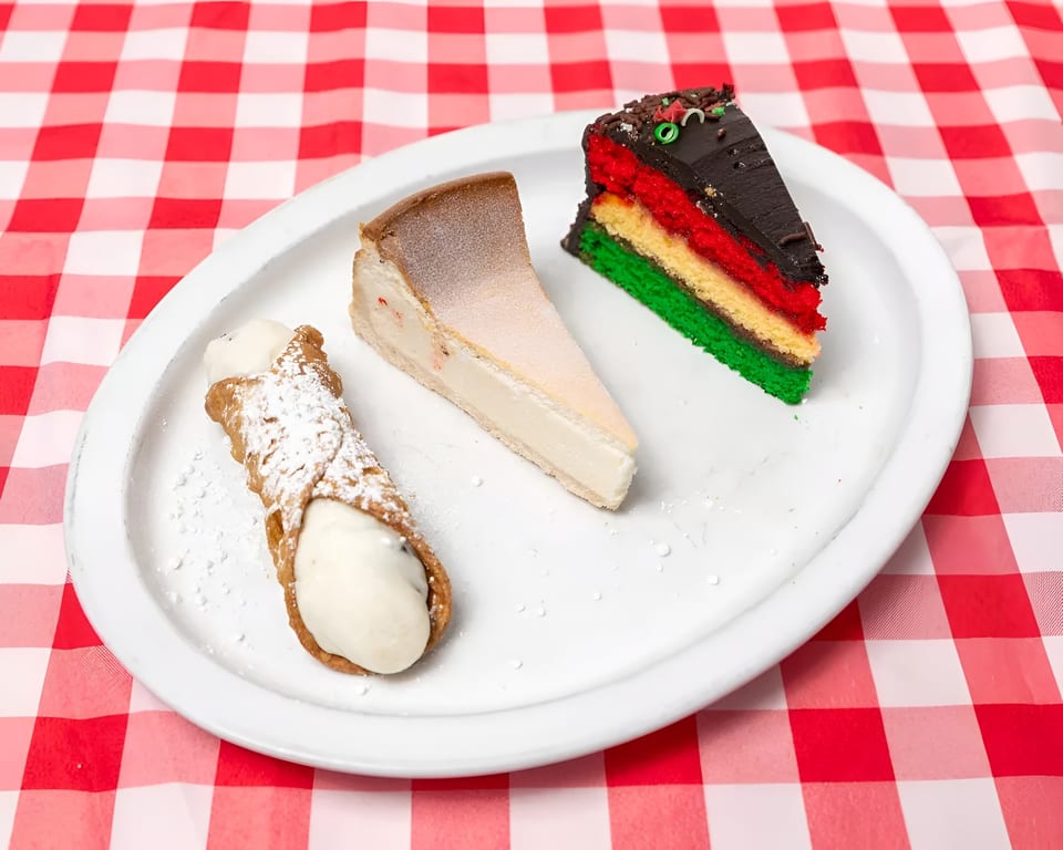 A cannoli, cheesecake slice, and rainbow cake slice on a white plate over a red and white checkered tablecloth.