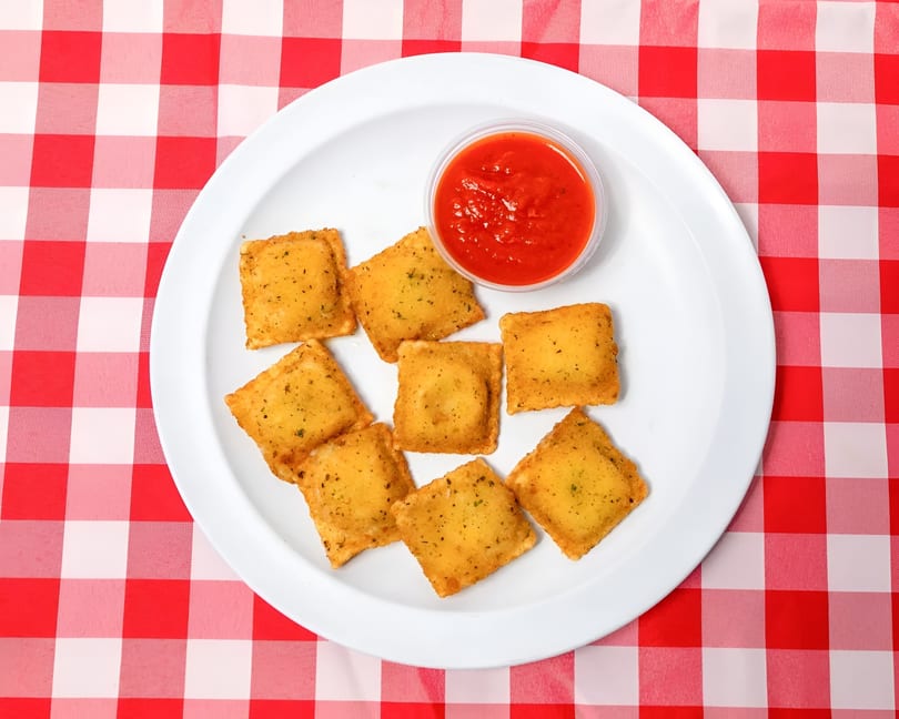 Golden fried ravioli with a side of red marinara sauce on a white plate over a red and white checkered tablecloth.