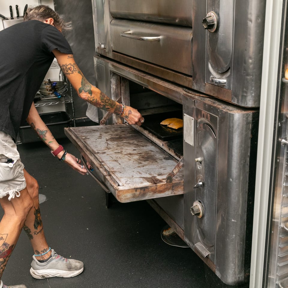 A person with tattooed arms and legs places flatbread into a large commercial oven.