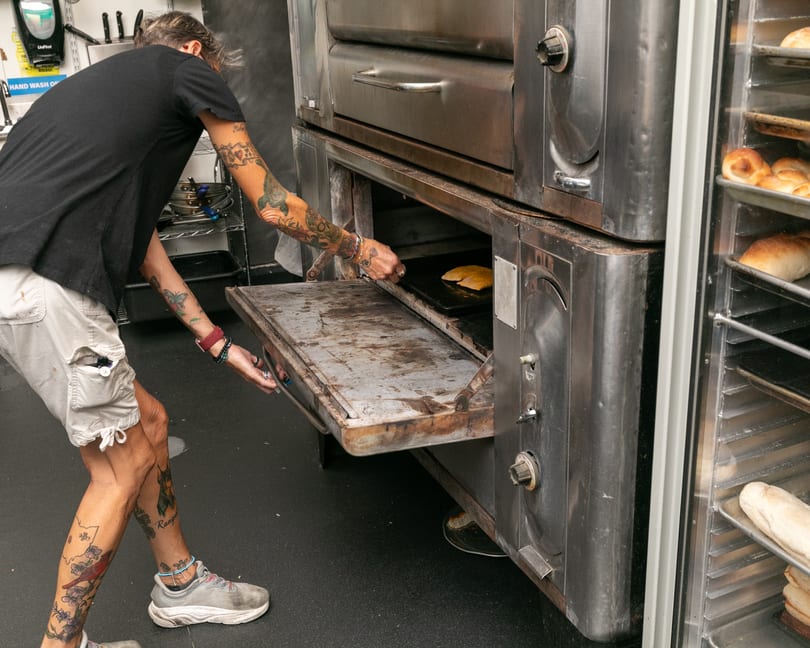A person with tattooed arms and legs places flatbread into a large commercial oven.