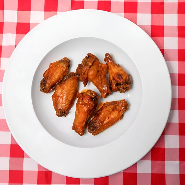 Six glazed chicken wings arranged on a white plate, set on a red and white checkered tablecloth.