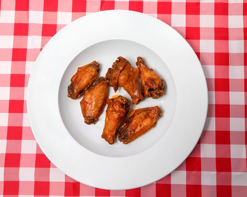 Six glazed chicken wings arranged on a white plate, set on a red and white checkered tablecloth.
