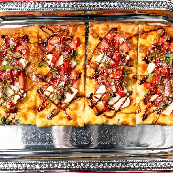 Rectangular flatbread pizza with tomatoes, mozzarella, basil, and balsamic glaze on a silver tray.