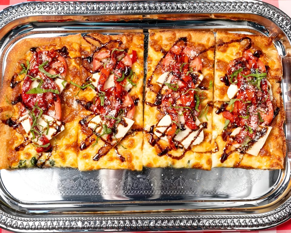 Rectangular flatbread pizza with tomatoes, mozzarella, basil, and balsamic glaze on a silver tray.