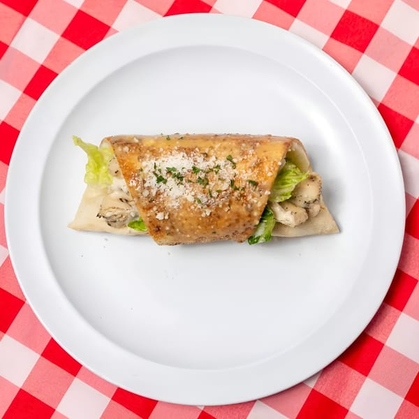 Chicken Caesar wrap with dressing, parmesan, and parsley on a white plate, on a red and white checkered tablecloth.