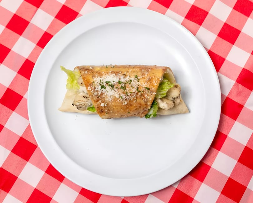 Chicken Caesar wrap with dressing, parmesan, and parsley on a white plate, on a red and white checkered tablecloth.