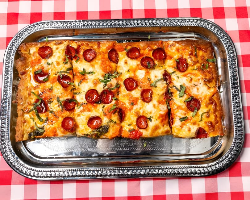 Rectangular pepperoni pizza with basil on a silver tray, on a red and white checkered tablecloth.