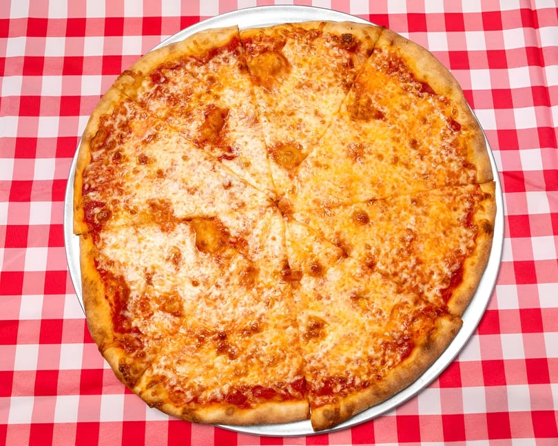A whole cheese pizza, cut into slices, on a metal tray over a red and white checkered tablecloth.