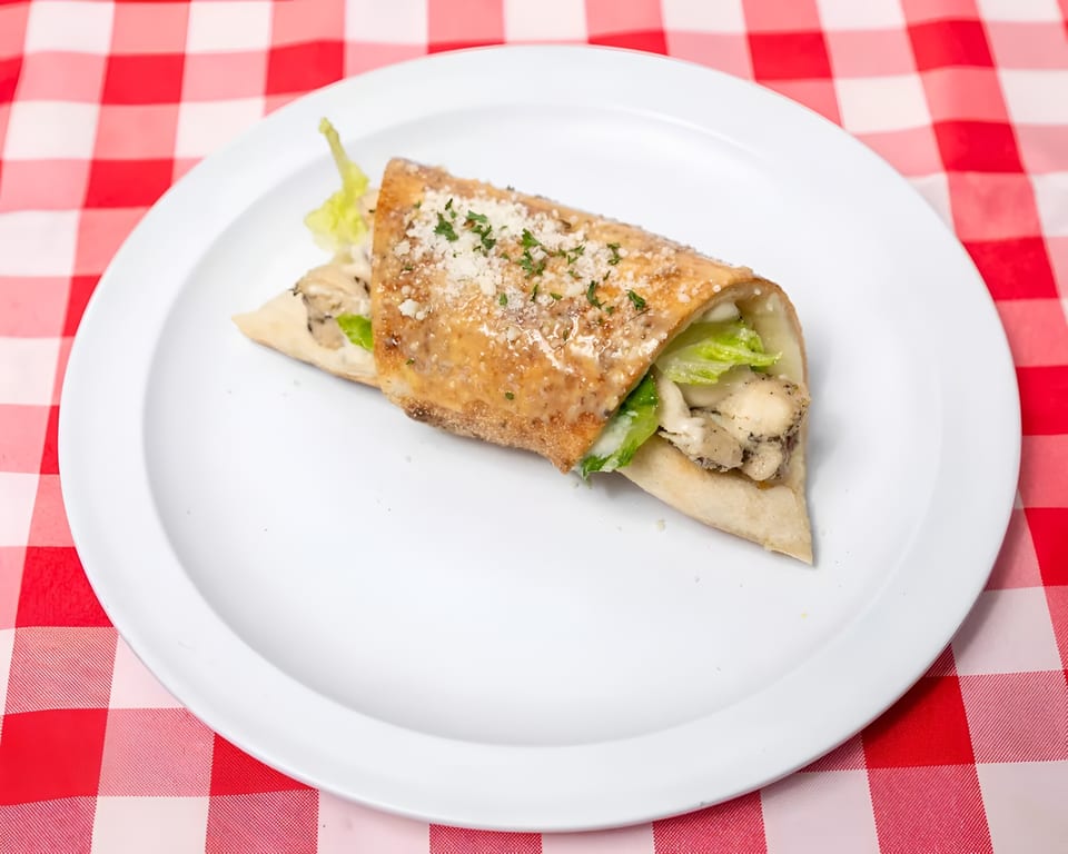 Chicken Caesar wrap with lettuce, parmesan, and dressing on a white plate over a red and white checkered tablecloth.