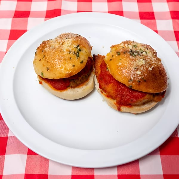 Two meatball sliders with red sauce and parmesan cheese on a white plate, on a red and white checkered tablecloth.