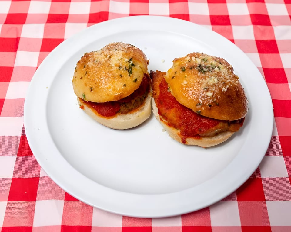 Two meatball sliders with red sauce and parmesan cheese on a white plate, on a red and white checkered tablecloth.