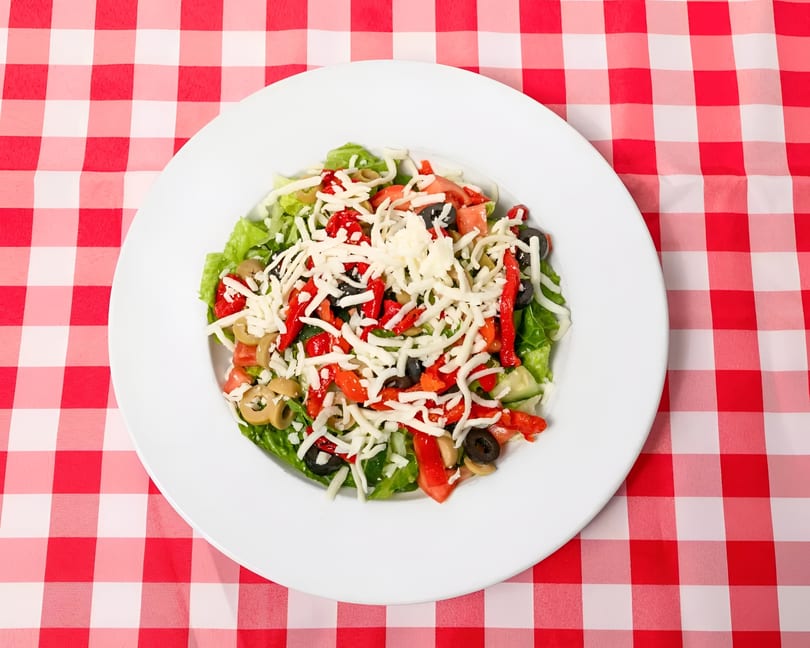 A fresh garden salad with lettuce, tomatoes, olives, red peppers, and shredded cheese on a red and white checkered tablecloth.